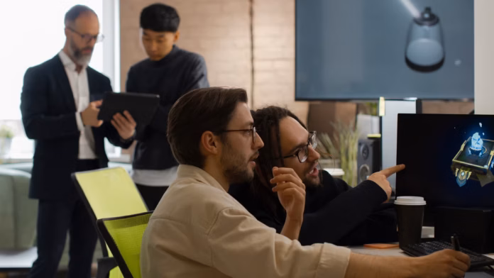 gamen.pro two game developers at a desk looking and pointing at a screen with two other game developers in the back blurred and looking at a tablet